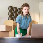 A young woman working on her laptop surrounded by cardboard boxes, indicating online business operations.